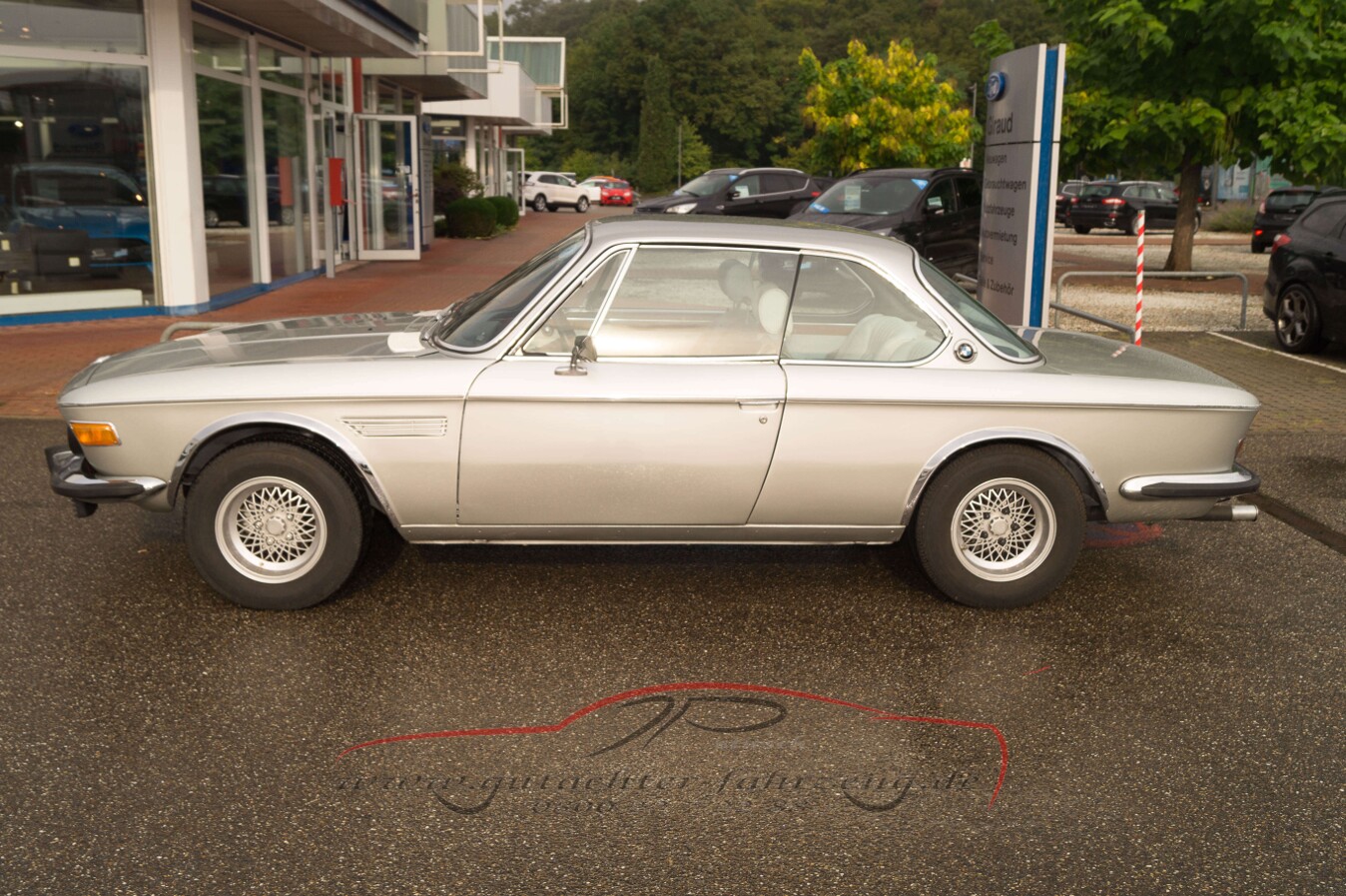 Ein silbernes klassisches Sportcoupé steht auf einem Parkplatz vor einem Autohaus.