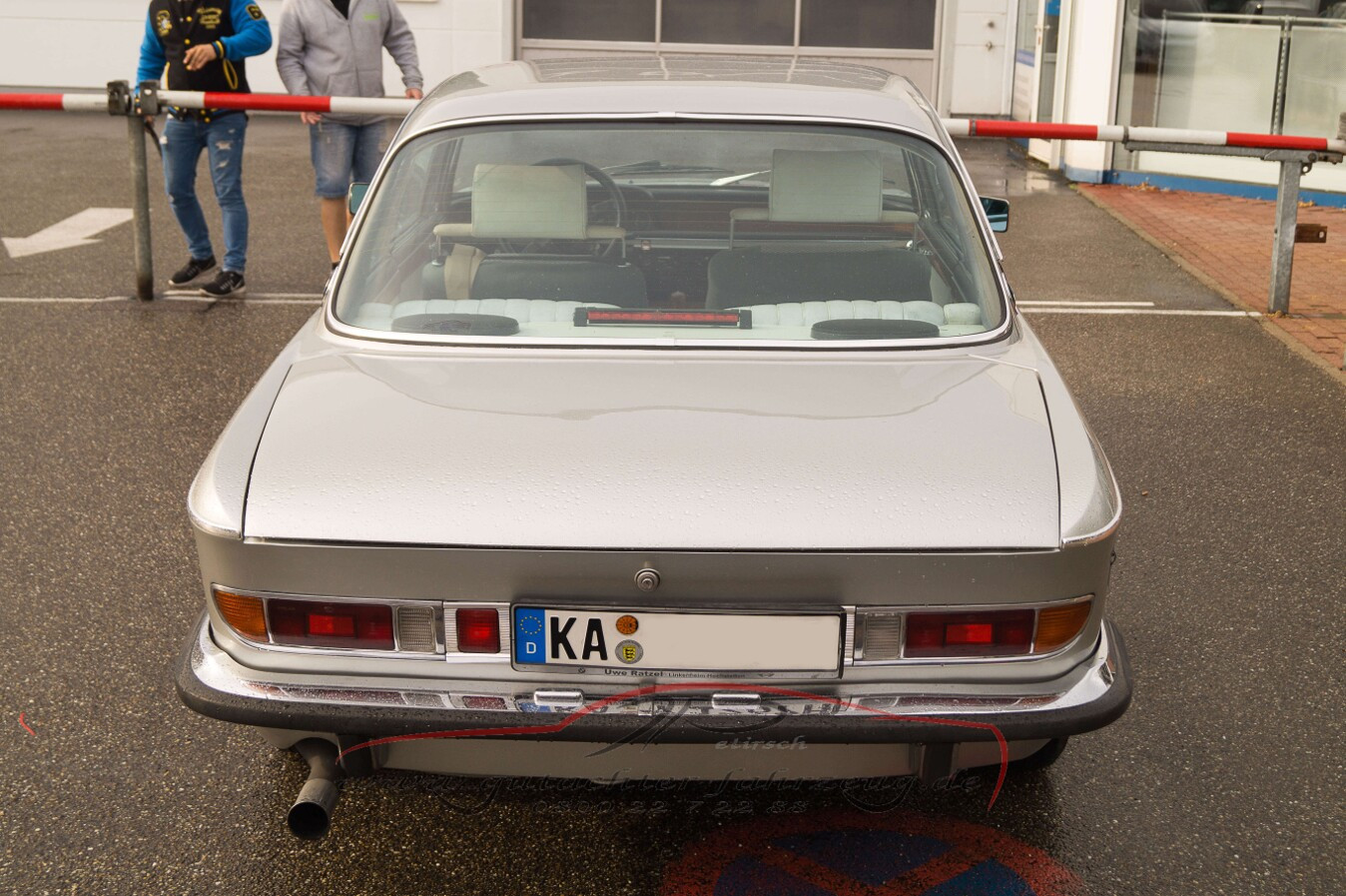 Ein silbernes Auto mit deutschem Kennzeichen steht vor einem geschlossenen Barrieretor, während zwei Personen im Hintergrund stehen.