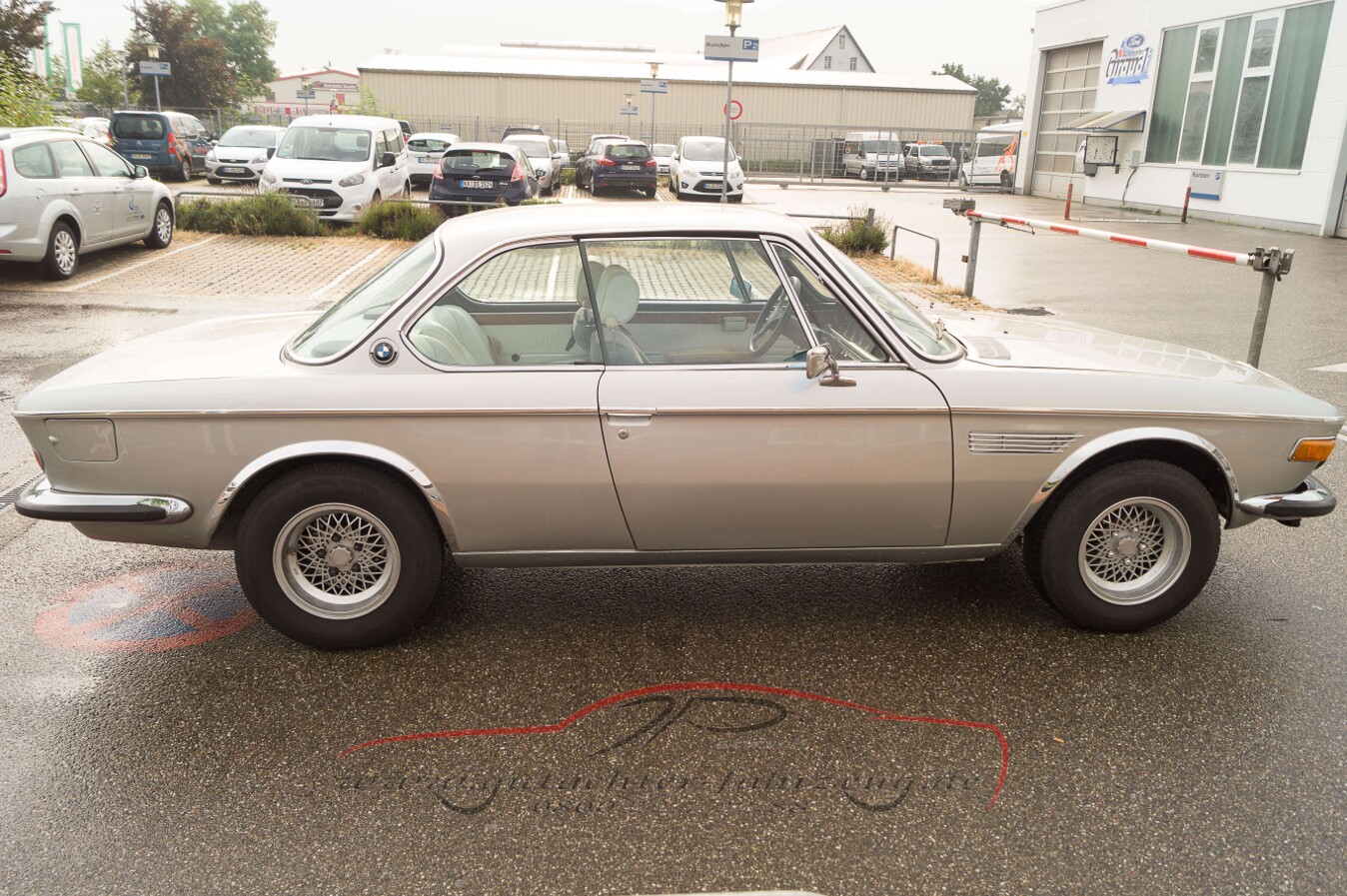 Ein silberfarbenes klassisches BMW-Coupé steht in einem Parkplatz vor einem Gebäude mit weiteren Autos im Hintergrund.