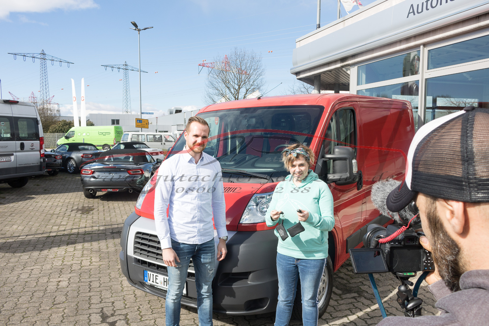 Zwei Personen stehen neben einem roten Lieferwagen in einem Autohauspark, während eine dritte Person mit einer Kamera filmt.