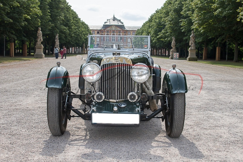 Ein grünes Vintage-Auto steht auf einer gepflasterten Allee vor einem Schloss mit Bäumen und Statuen.
