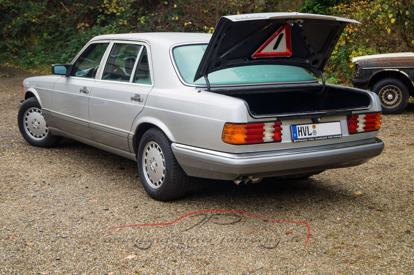 Ein silbernes Auto mit geöffneter Kofferraumklappe steht auf einem Kiesweg, im Hintergrund sind Bäume und ein weiteres Fahrzeug zu sehen.