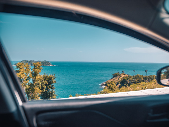 Blick aus einem Autofenster auf ein türkisfarbenes Meer mit einer Insel im Hintergrund und grünen Bäumen am Rand.
