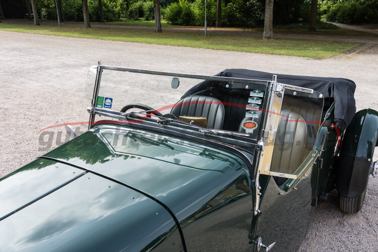 Ein grünes Oldtimer-Auto mit offener Kabine und einem klaren Windschutzschild steht auf einem Parkplatz.