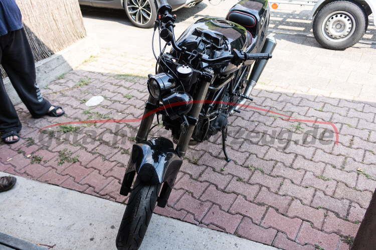 Ein schwarzes Motorrad steht auf einem gepflasterten Gehweg, im Hintergrund sind Autos und Teile von Personen zu sehen.