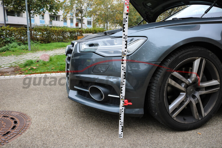 Ein graues Auto mit geöffnetem Kofferraum steht auf einem Parkplatz, neben ihm steht ein Maßstab.