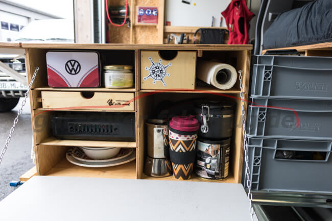Ein organisiertes Regal mit verschiedenen Gegenständen wie Tassen, Behältern und einem VW-Logo im Hintergrund.